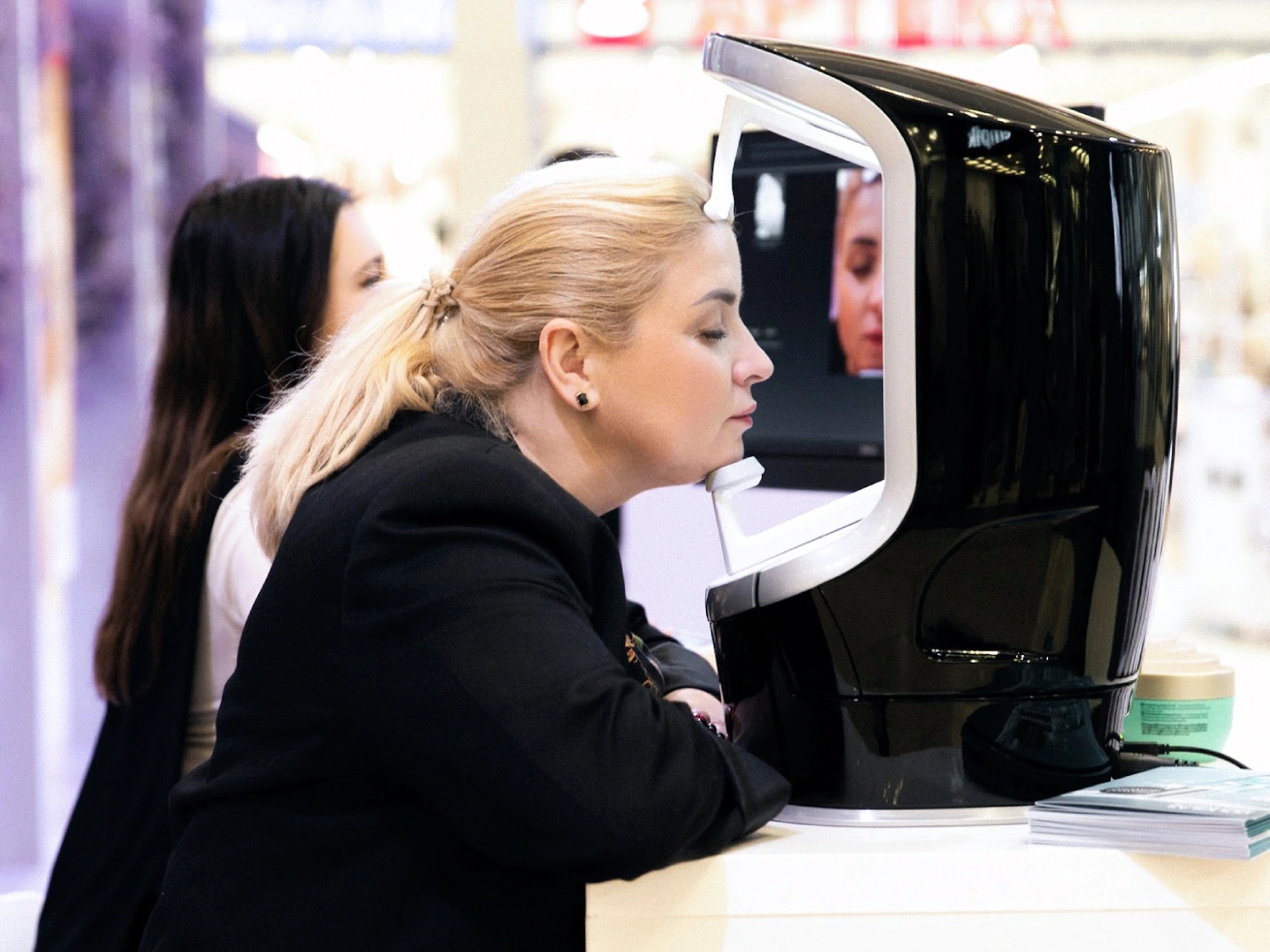 Kobieta z blond włosami spiętymi w kucyk poddaje się analizie skóry twarzy za pomocą specjalistycznego urządzenia diagnostycznego.