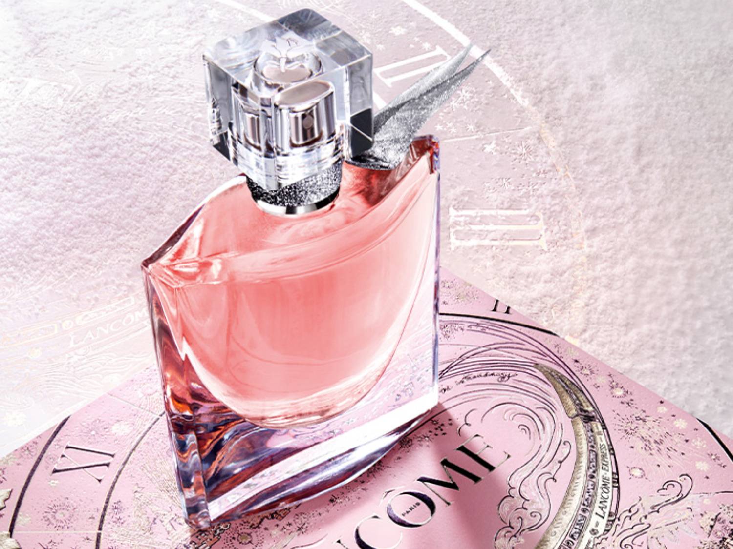 Różowy flakon perfum Lancôme La Vie Est Belle z przezroczystą nakrętką w kształcie kostki, umieszczony na ozdobnym, różowym pudełku z motywem zegara.
