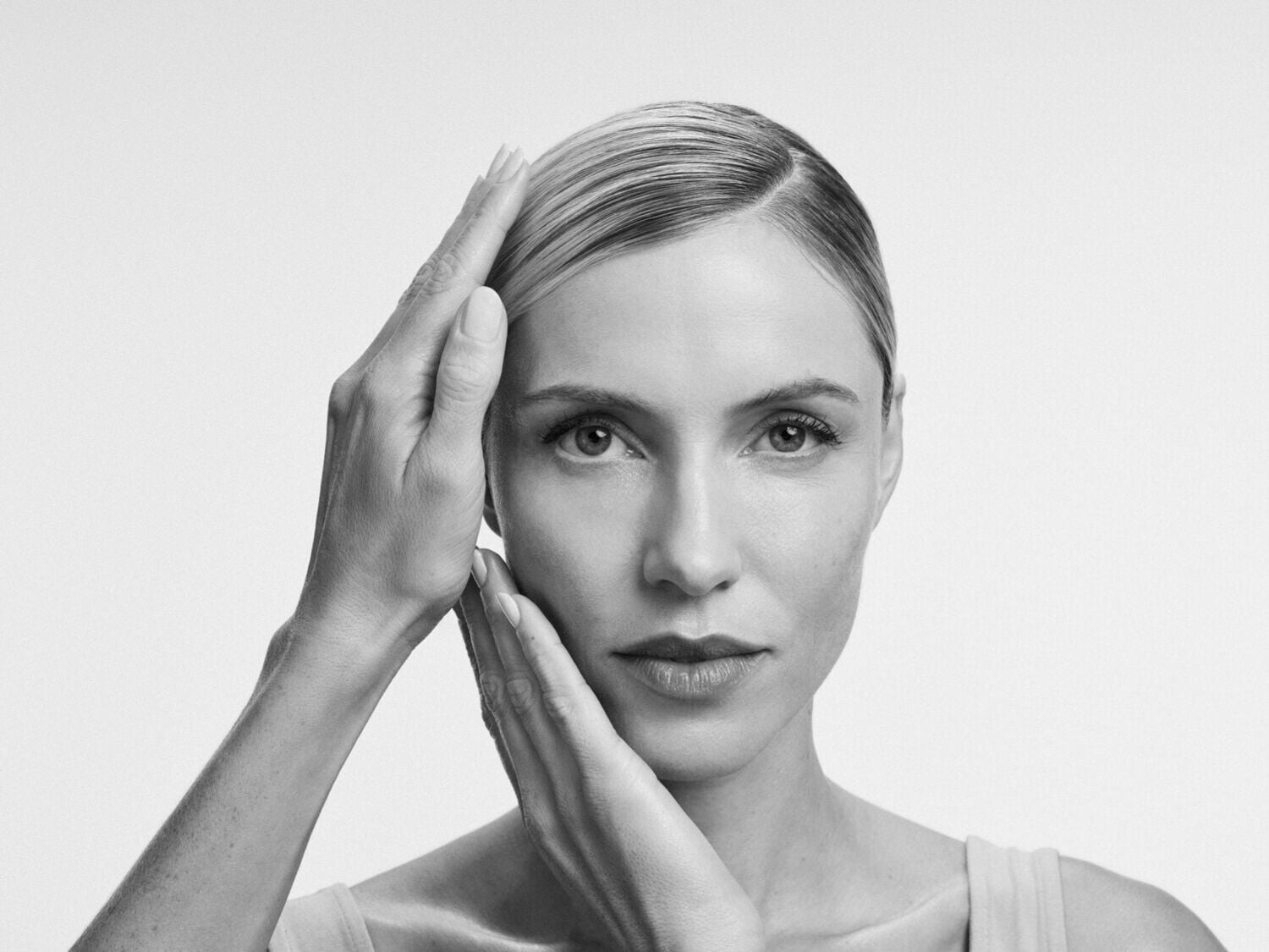 Moters portretas juodai baltas, rankos šalia veido, pabrėžiantis odos priežiūrą ir grožio rutiną.