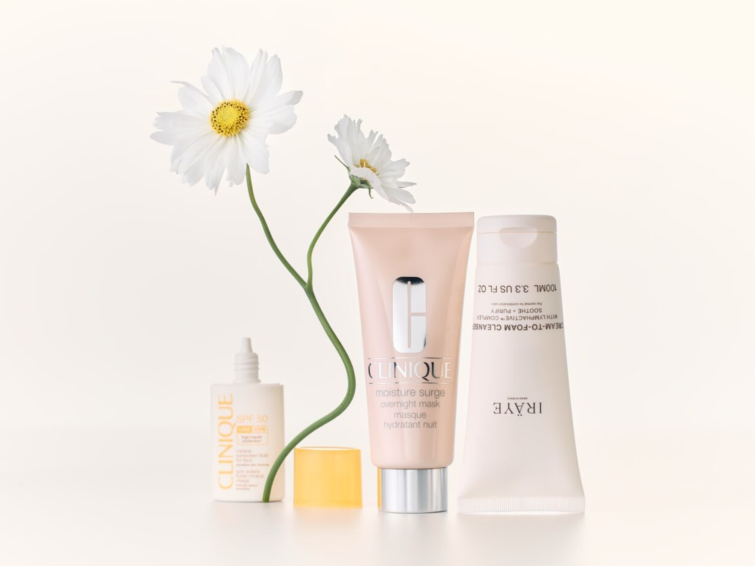 Weiße Gänseblümchen neben Clinique SPF 50, Moisture Surge Overnight Mask in rosa Tube und IRÄYE Cream-To-Foam Cleanser in weißer Tube.