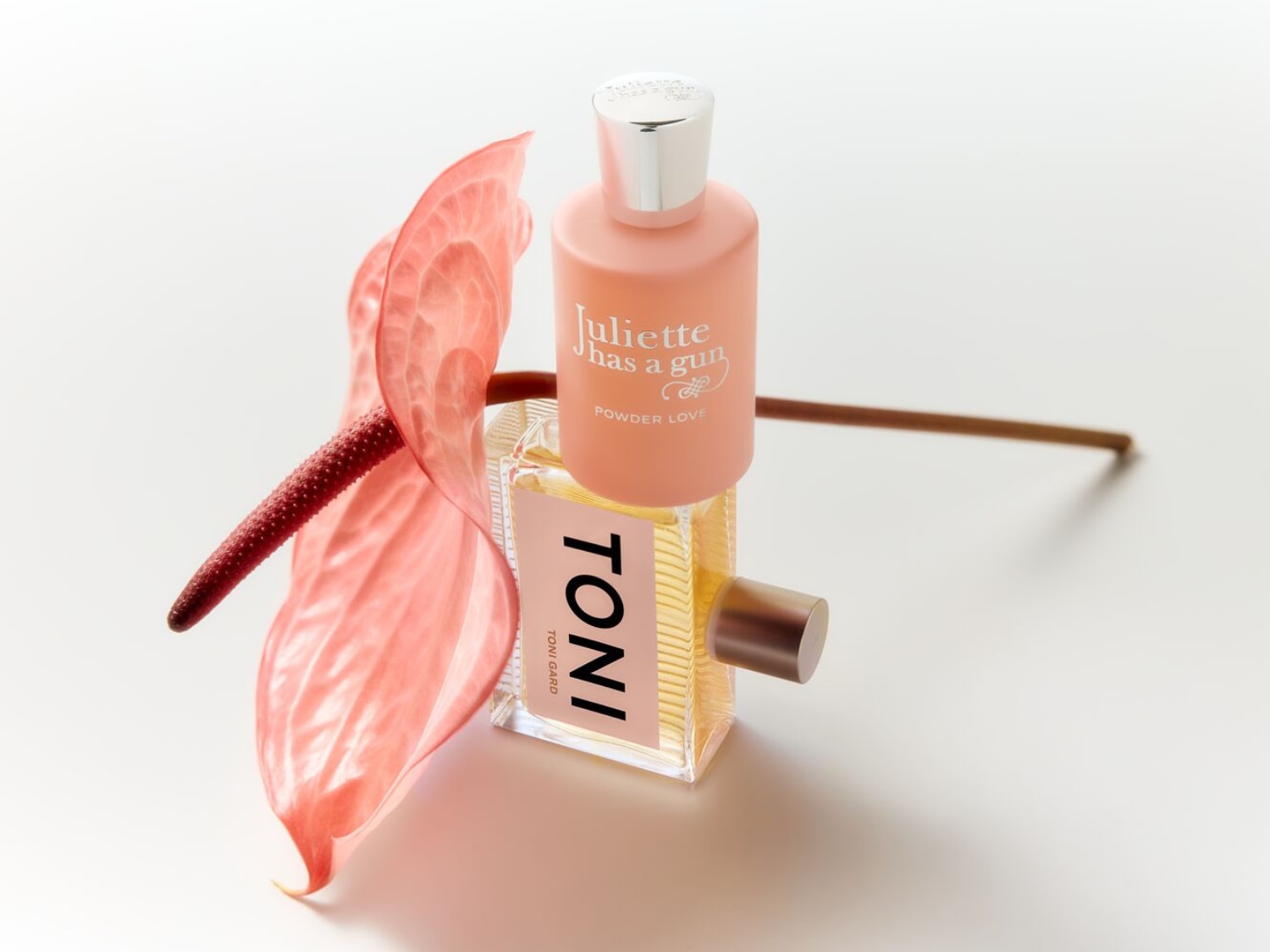 Ein Arrangement aus zwei Parfümflaschen, 'Juliette Has a Gun' in einem zarten Rosa und 'Toni Gard' mit einem Hauch von Gold, ergänzt durch eine elegante, rosa Anthurie.