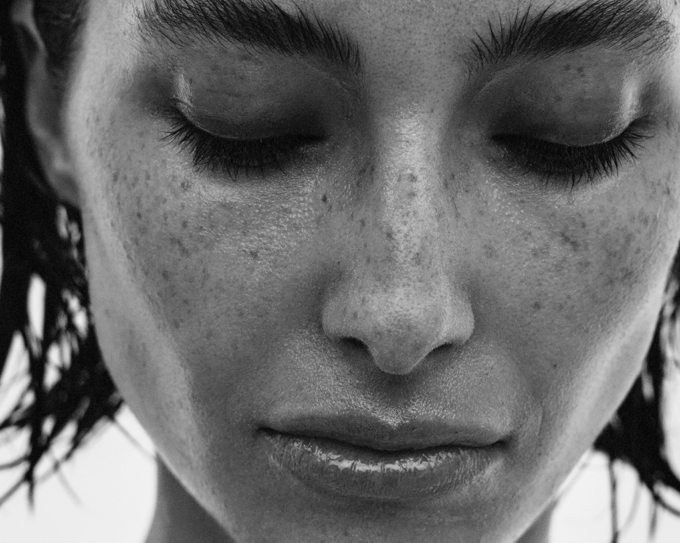 Czarno-białe zbliżenie twarzy kobiety z zamkniętymi oczami, pokazujące jej piegi i naturalny blask skóry.