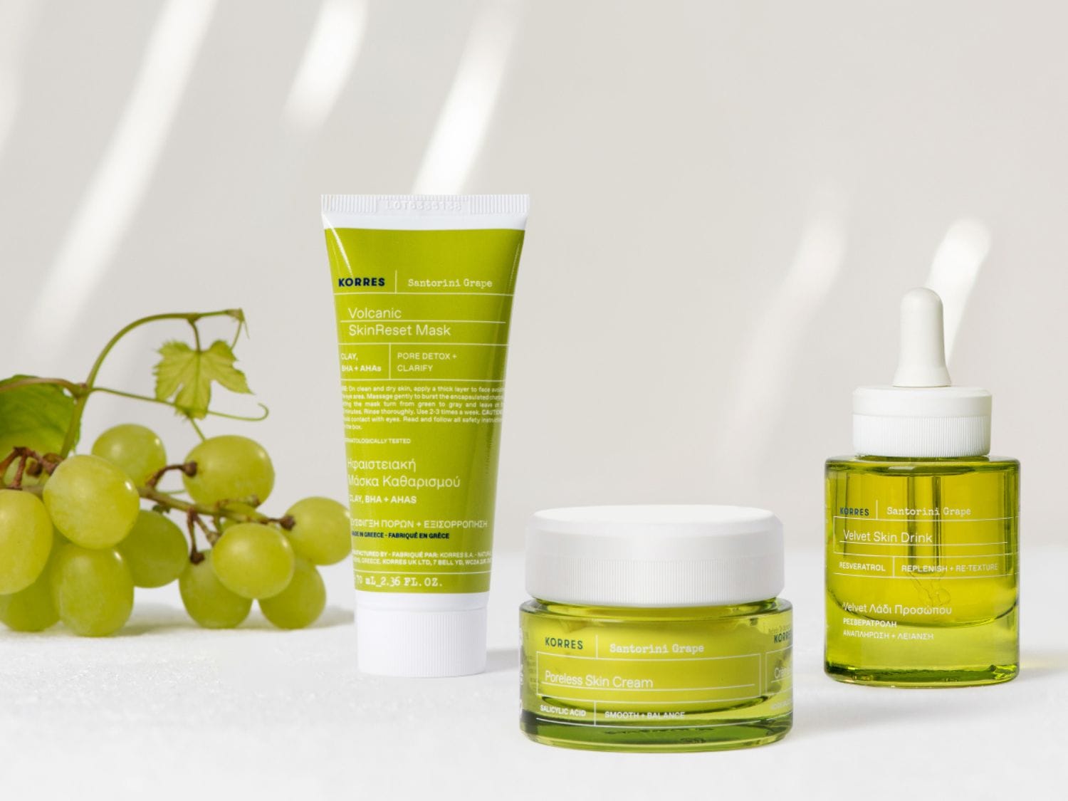 Na białym tle znajdują się trzy produkty do pielęgnacji skóry marki Korres Santorini Grape w zielonych opakowaniach oraz kiść zielonych winogron.