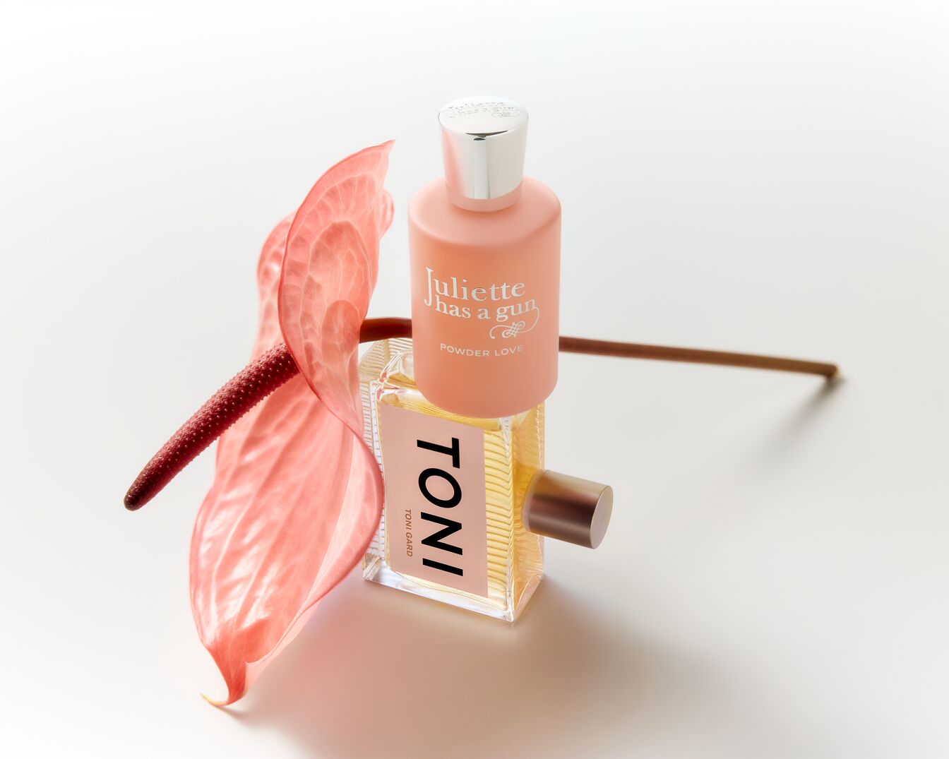 Ein Arrangement zeigt zwei Parfümflaschen, eine in zartem Rosa mit der Aufschrift 'Juliette has a gun POWDER LOVE' und eine goldfarbene Flüssigkeit enthält, neben einer rosa Anthurie.