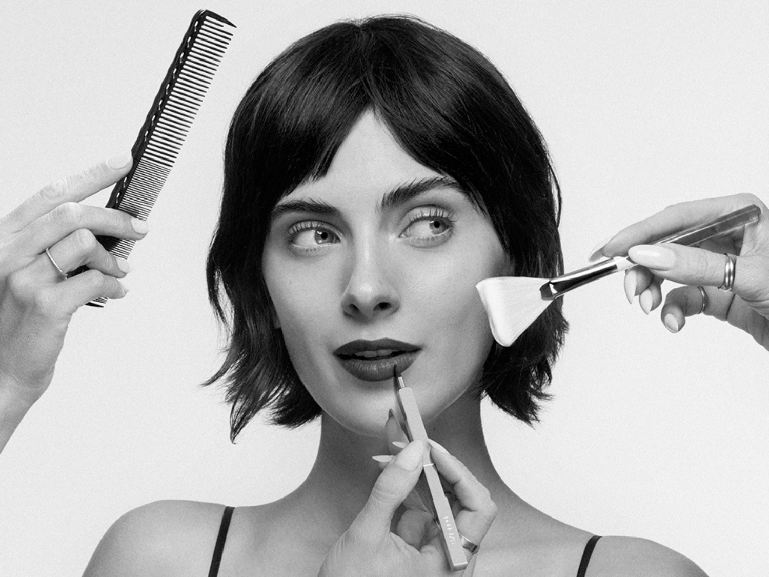 Eine Frau mit dunklen Haaren wird mit einem Kamm frisiert, während ein Pinsel eine Maske aufträgt und ein Lippenstift die Lippen betont.