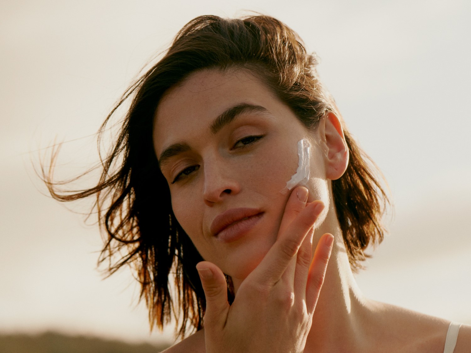 Pohľad zblízka na ženu, ktorá si nanáša biely krém na tvár, pričom jej hnedé vlasy vejú vo vánku.