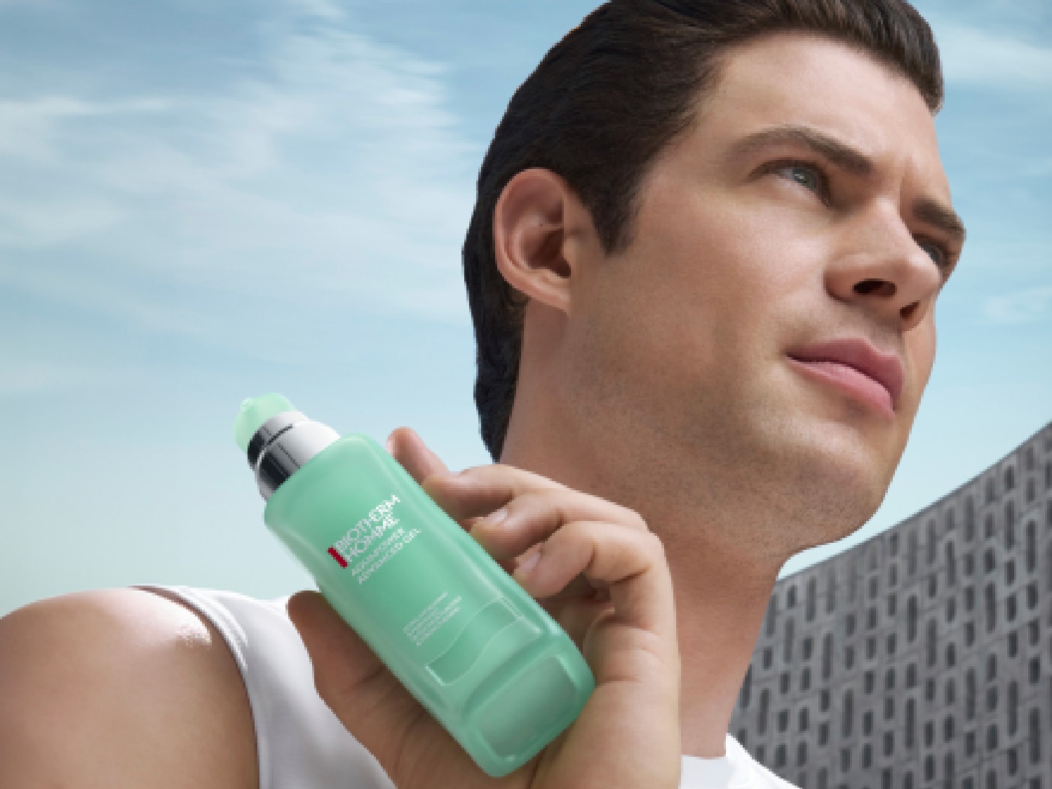 Un homme tient un flacon vert d'Aquapower Advanced Gel de Biotherm Homme, avec un ciel bleu en arrière-plan, pour une peau hydratée et revitalisée.