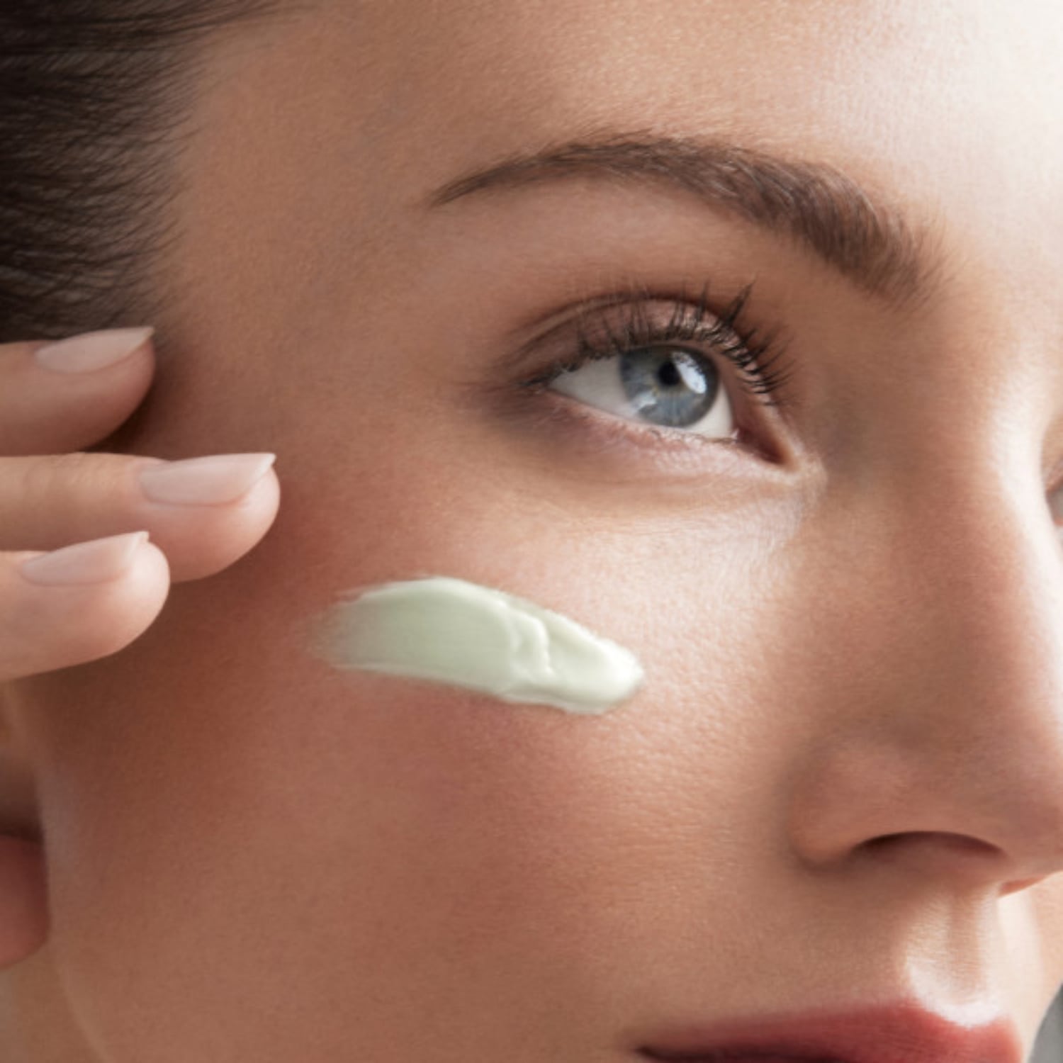 Gros plan sur une femme appliquant une crème verte sur sa joue, mettant en valeur sa peau et son regard clair.
