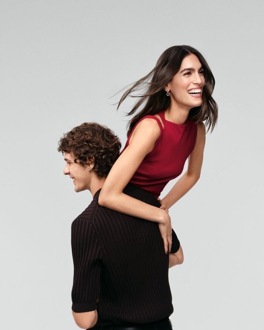 Un homme portant une femme sur son dos, tous deux souriant et regardant vers le haut, elle porte un haut rouge sans manches et il porte un polo noir.