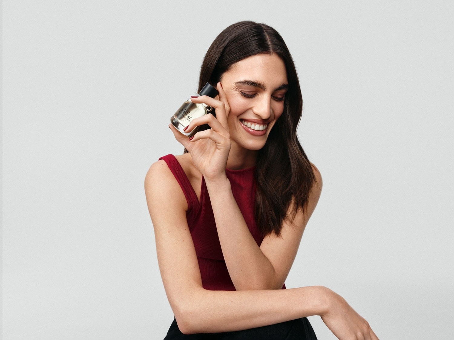 Une femme souriante tient un flacon de parfum transparent avec un bouchon noir, portant un haut rouge et un pantalon noir.