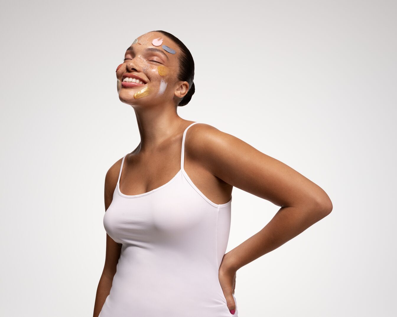 Una donna sorridente con diverse maschere colorate applicate sul viso, indossa una canotta bianca e ha i capelli raccolti.