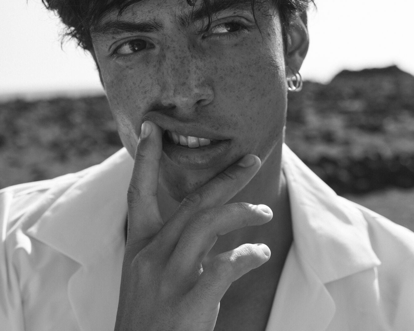 Schwarz-Weiß-Porträt eines Mannes mit Sommersprossen, der ein weißes Hemd trägt und einen silbernen Ohrring, während er nachdenklich mit dem Finger an der Lippe spielt.