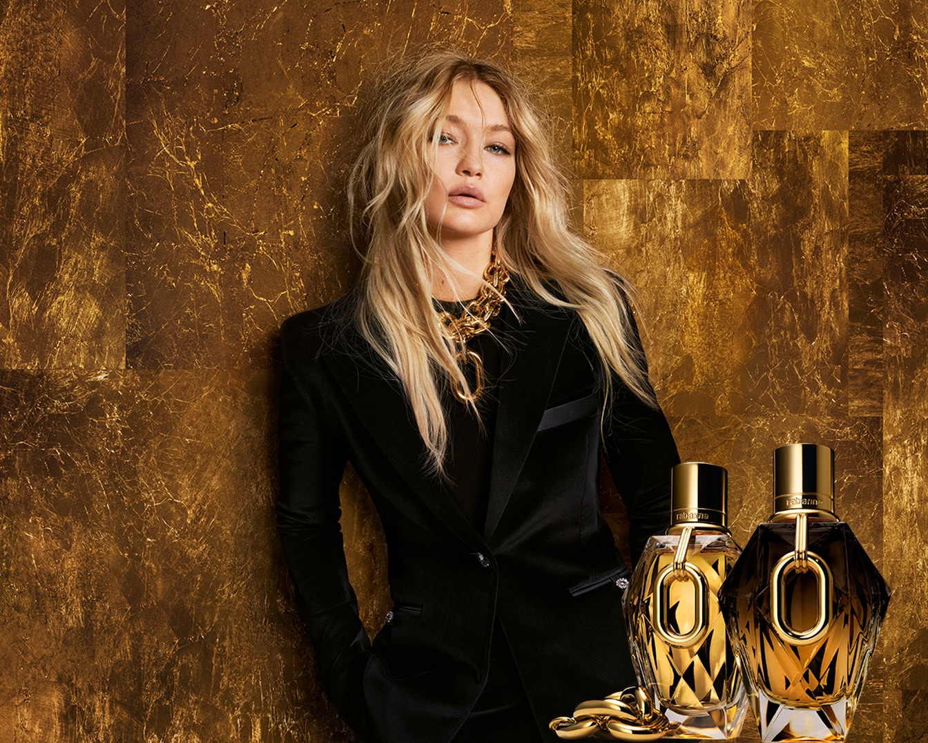 Blonde Frau mit Goldkette und schwarzem Blazer posiert neben zwei goldenen Parfümflaschen vor einem goldenen Hintergrund.