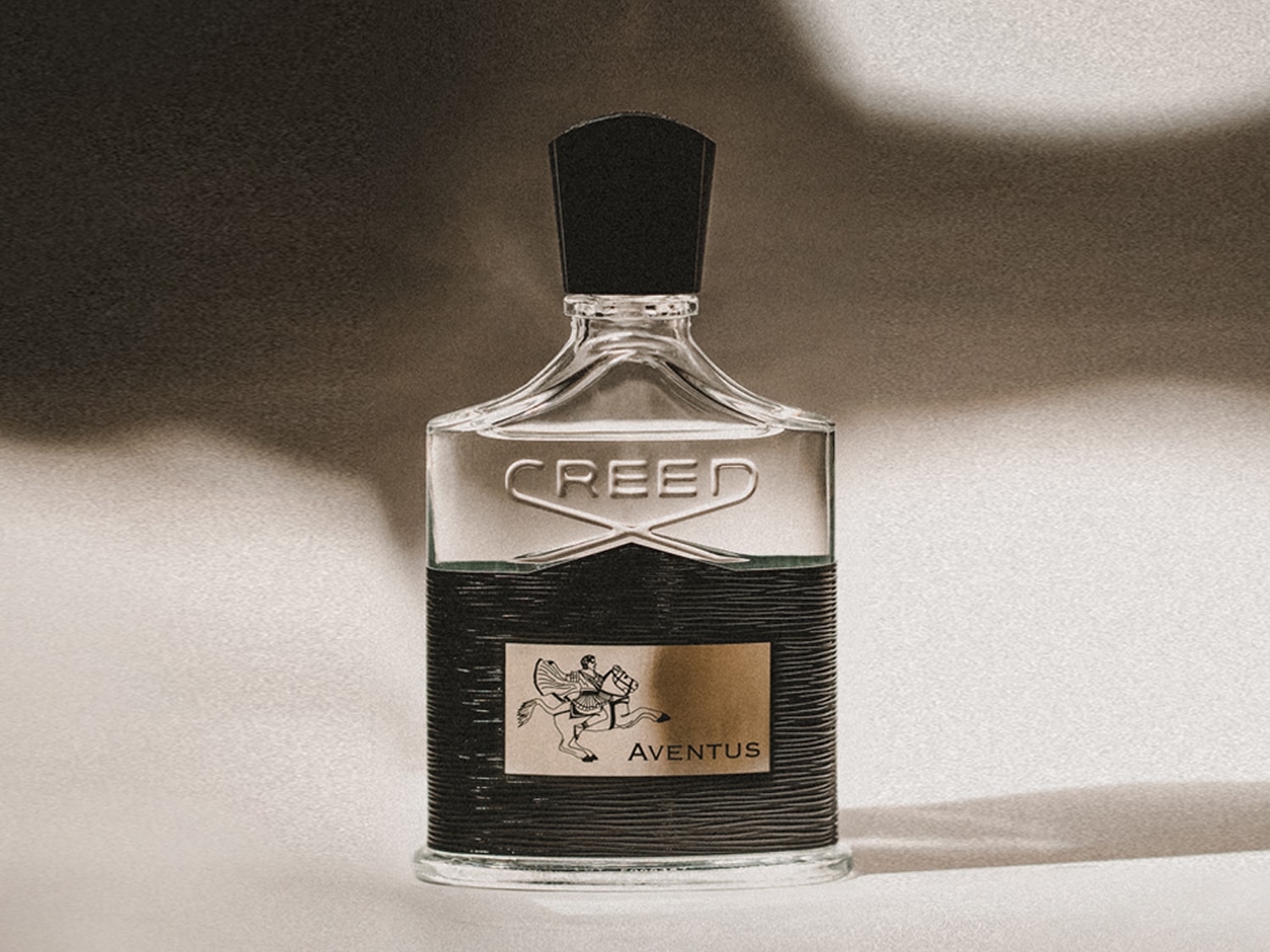 A Creed Aventus parfümös üveg átlátszó, fekete kupakkal, az üveg alsó részén fekete textúrával és arany címkével, amelyen egy lovas látható.