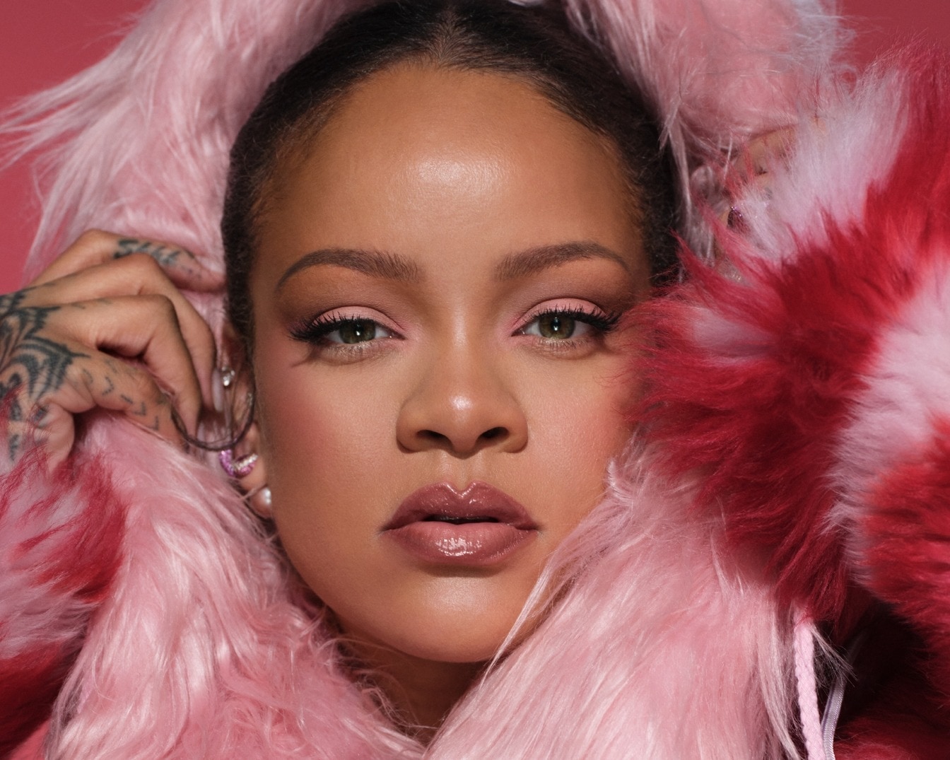 Close-up van Rihanna met een zachte roze make-up en een pluizige roze en rode jas, haar blik recht in de camera.