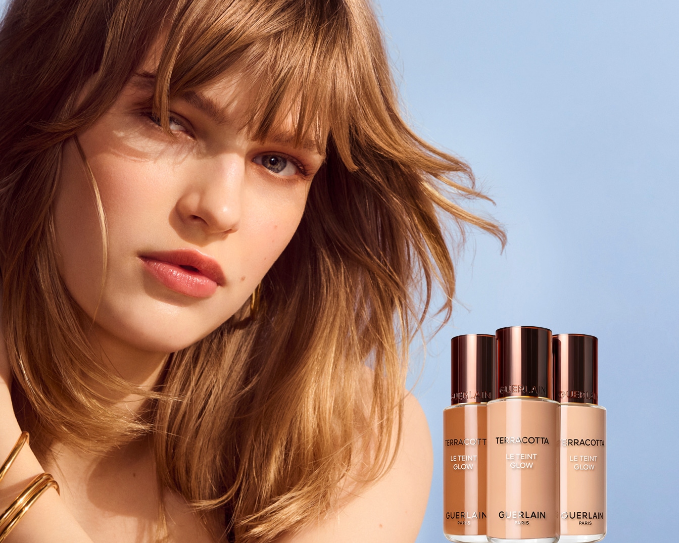 Na obrázku je žena s hnedými vlasmi a tromi fľaštičkami make-upu Terracotta Le Teint Glow od Guerlain, ktoré sľubujú žiarivý vzhľad.
