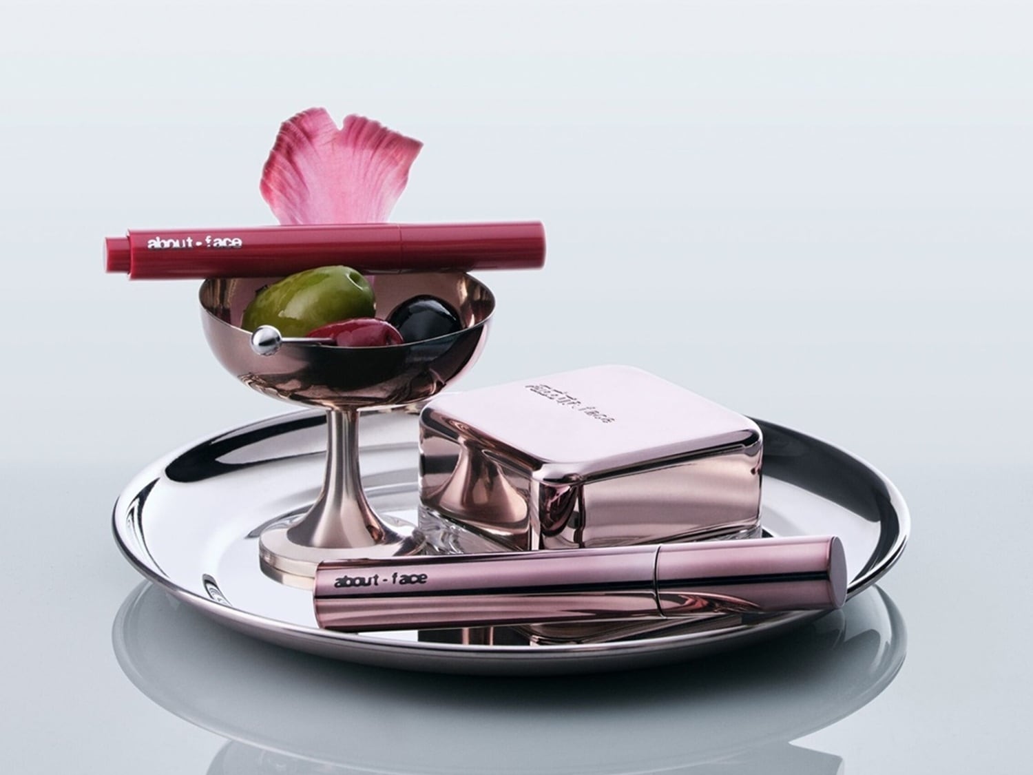 Auf einem silbernen Tablett sind Make-up-Produkte von About-Face in Roségold drapiert, darunter ein Lippenstift auf einer Schale mit Früchten und Blütenblatt.