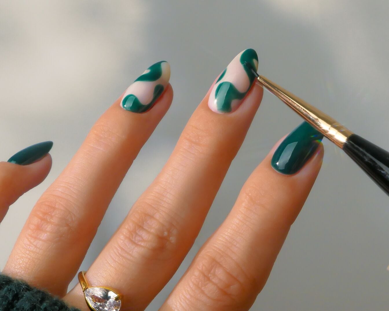 Nahaufnahme einer Hand, die mit einem Pinsel grüne Farbe auf einen elfenbeinfarbenen Nagel aufträgt, um ein einzigartiges Design zu kreieren.