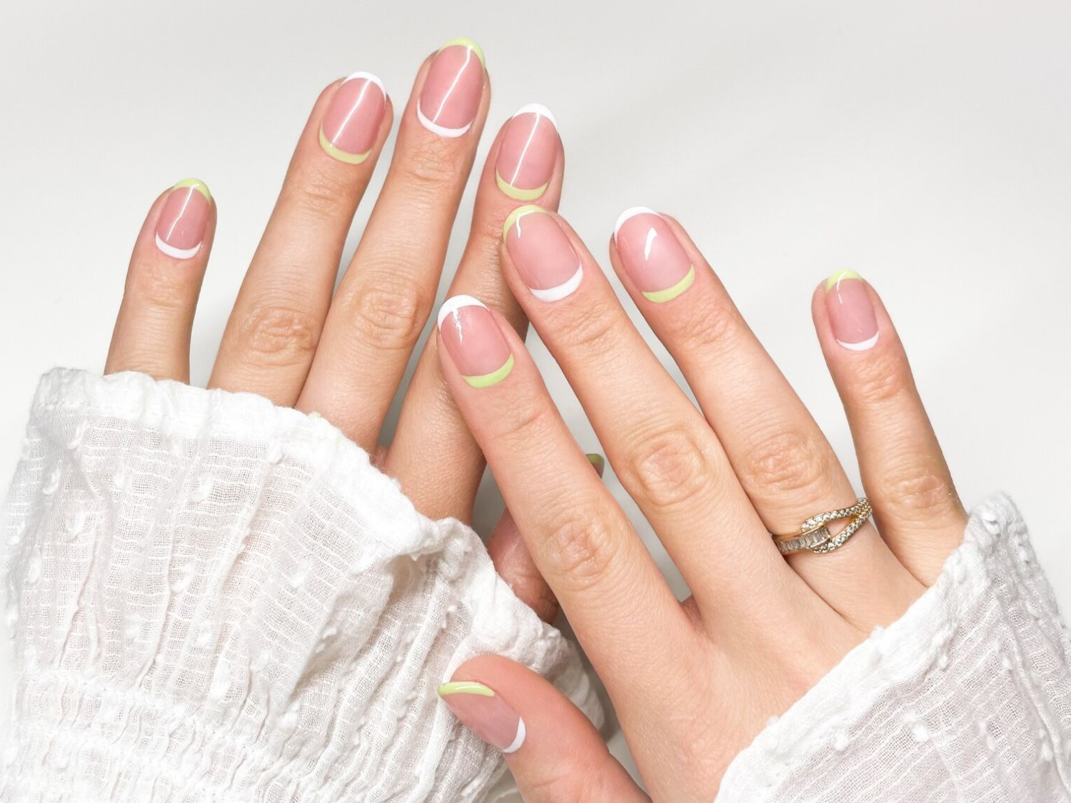 Nahaufnahme von zwei Händen mit mandelförmigen Nägeln, die mit einem weißen und lindgrünen French-Tip-Design verziert sind, und einem goldenen Ring am Finger.