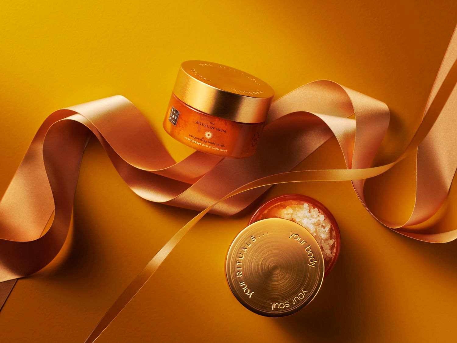 Das Ritual von Mehr belebendes Körperpeeling mit süßer Orangenschale und Zedernholz in goldener Verpackung, umgeben von einer goldenen Schleife.