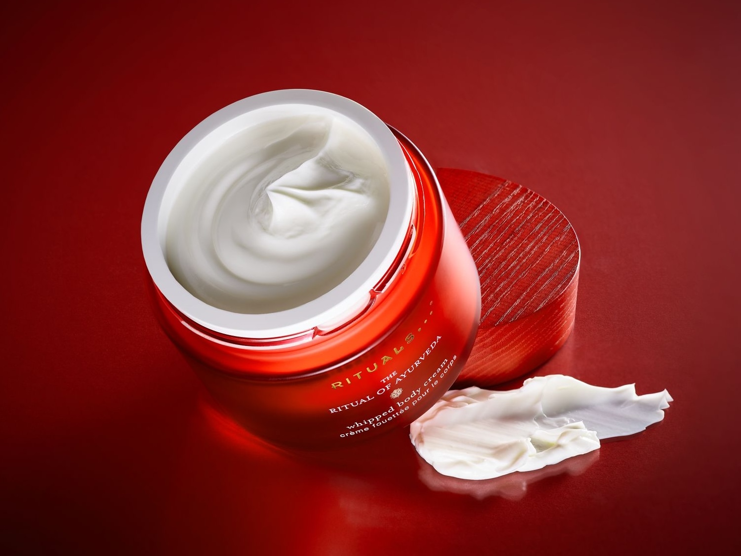 Ein offener, orangeroter Tiegel Rituals The Ritual of Ayurveda Whipped Body Cream mit weißer Creme und danebenliegendem Deckel auf rotem Grund.