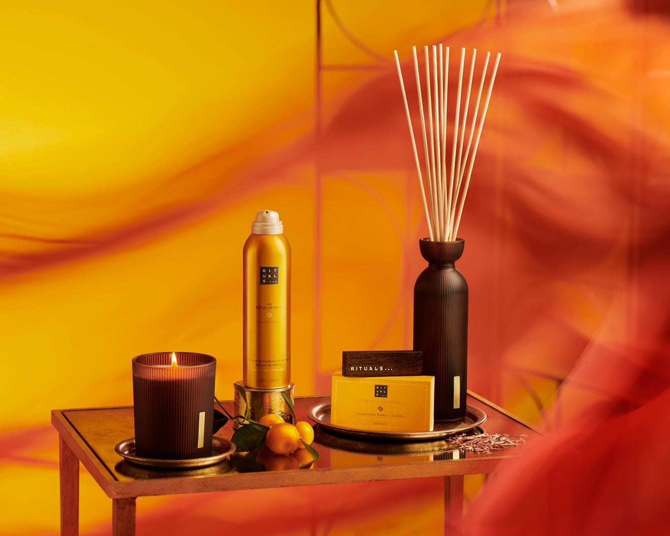 Auf einem goldenen Tisch stehen eine Duftkerze, ein Raumspray in Gold, Duftstäbchen in einer dunklen Vase und eine gelbe Schachtel von Rituals.