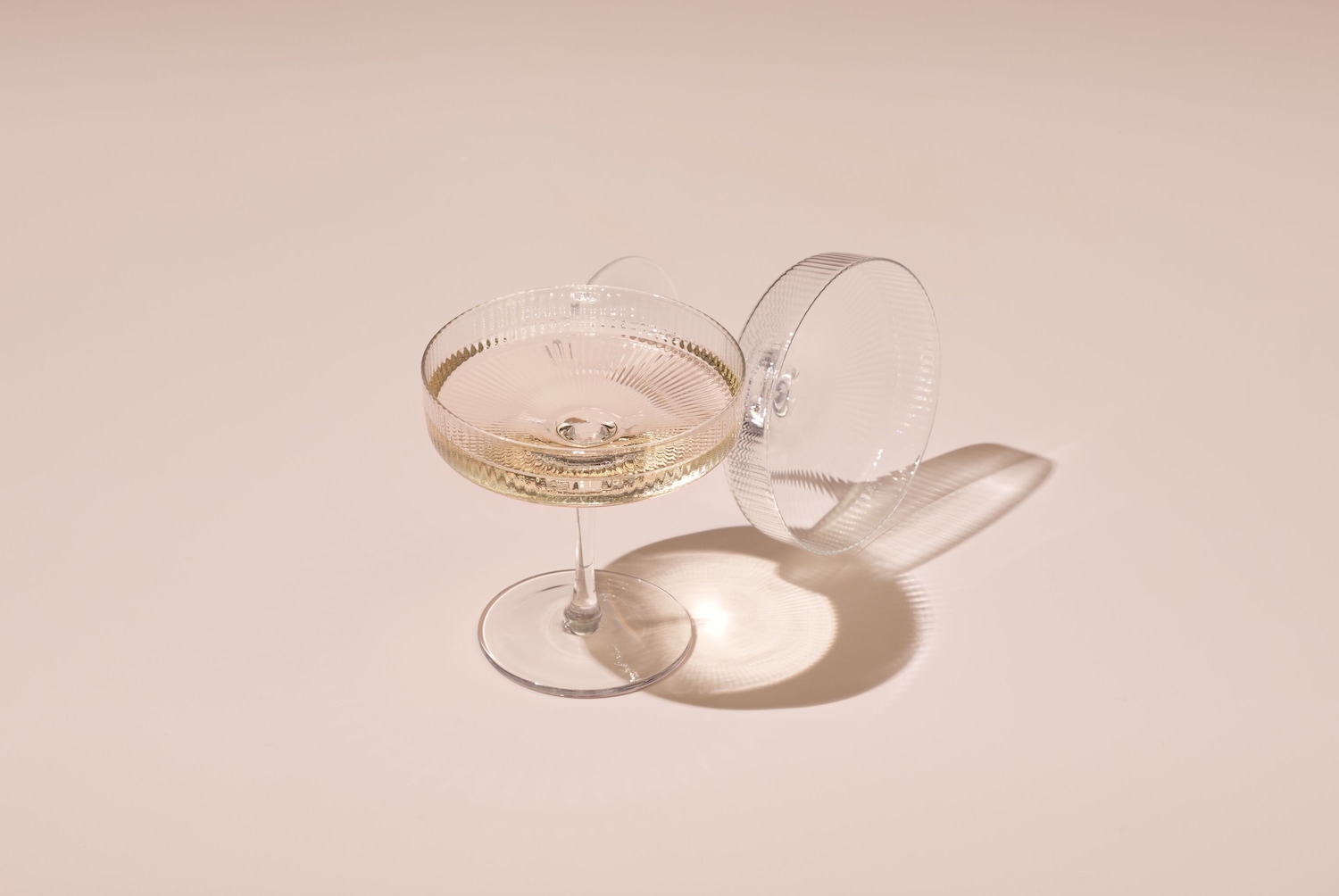 Auf einem hellrosa Hintergrund stehen zwei elegante, durchsichtige Cocktailgläser, eines gefüllt mit einem hellen Getränk, das andere liegt daneben.