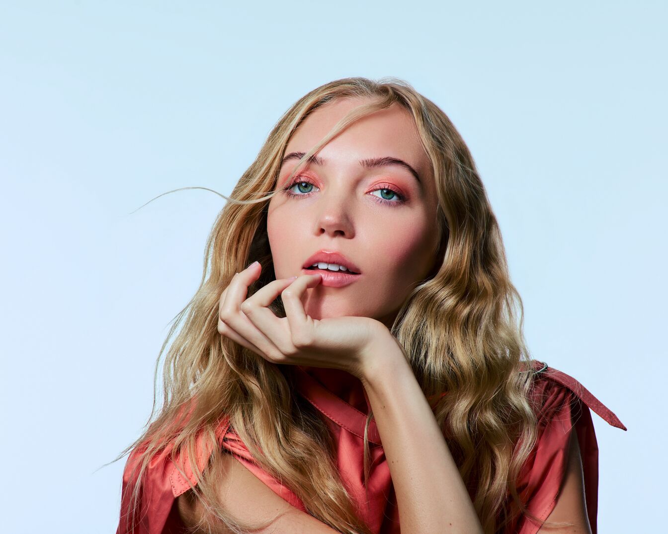 Een jonge vrouw met lang blond haar, gekleed in een roze top, kijkt omhoog met een hand onder haar kin, haar lippen lichtjes geopend en haar ogen opgemaakt met roze oogschaduw.