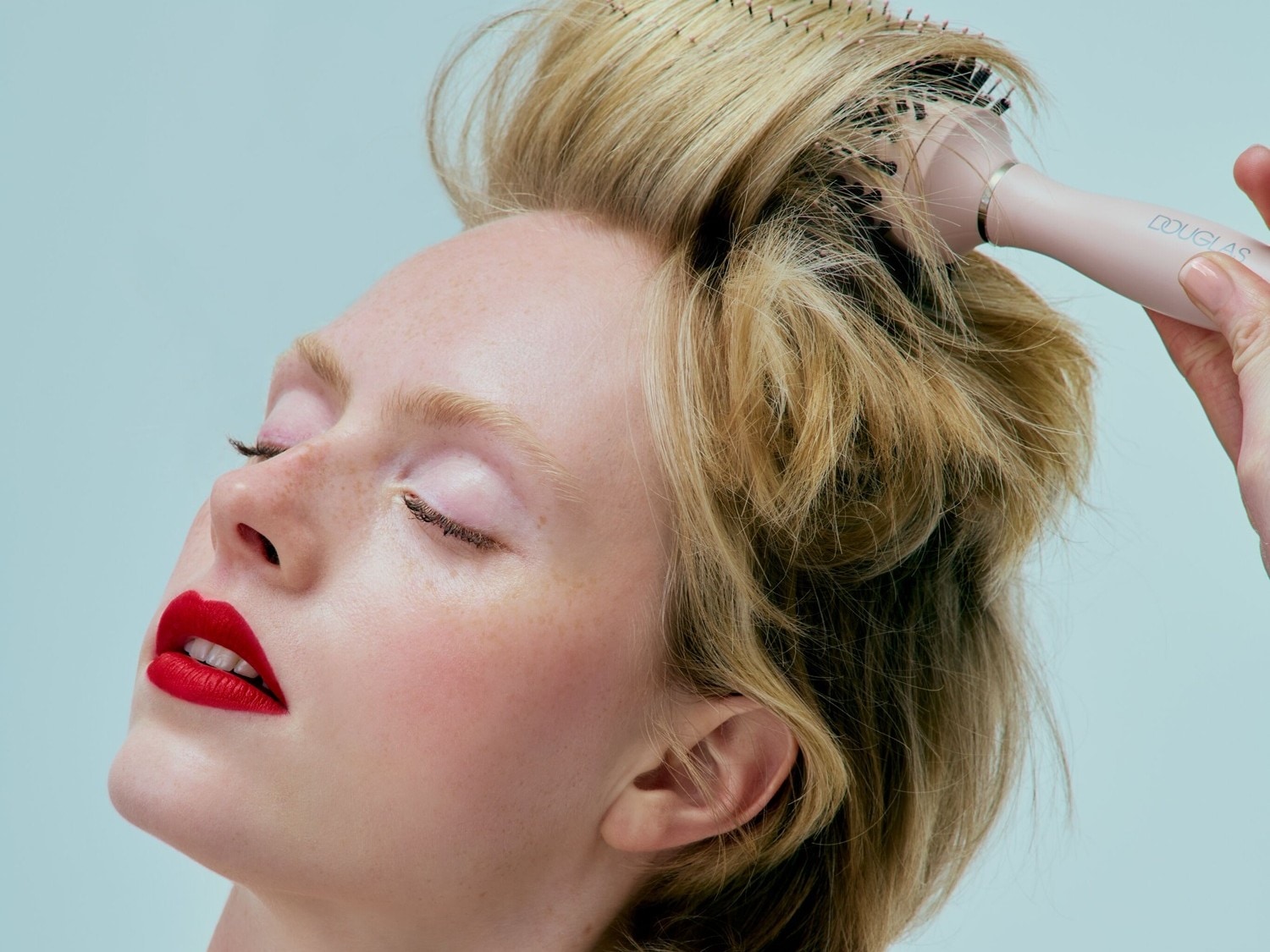 Eine blonde Frau mit geschlossenen Augen und roten Lippen wird mit einer rosafarbenen Rundbürste von Douglas frisiert.