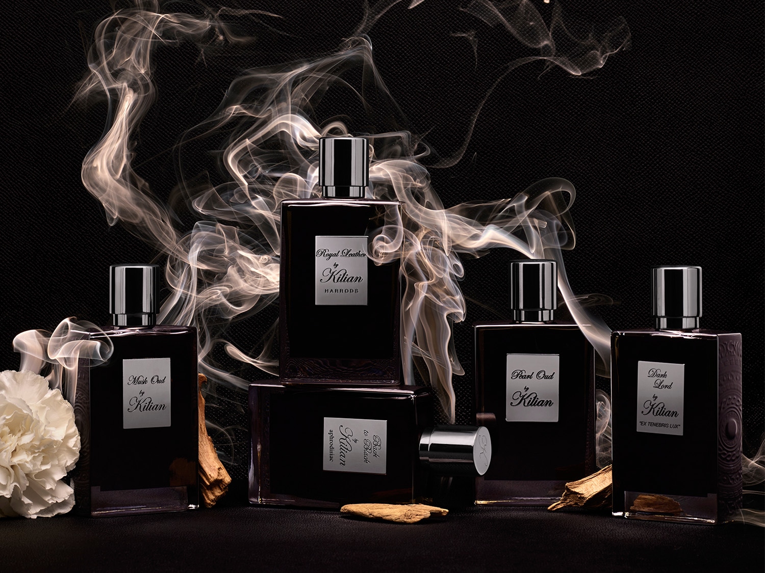 Eine Sammlung von Kilian-Parfums in dunklen Flakons mit silbernen Etiketten, umgeben von Rauchschwaden und einer Blüte.