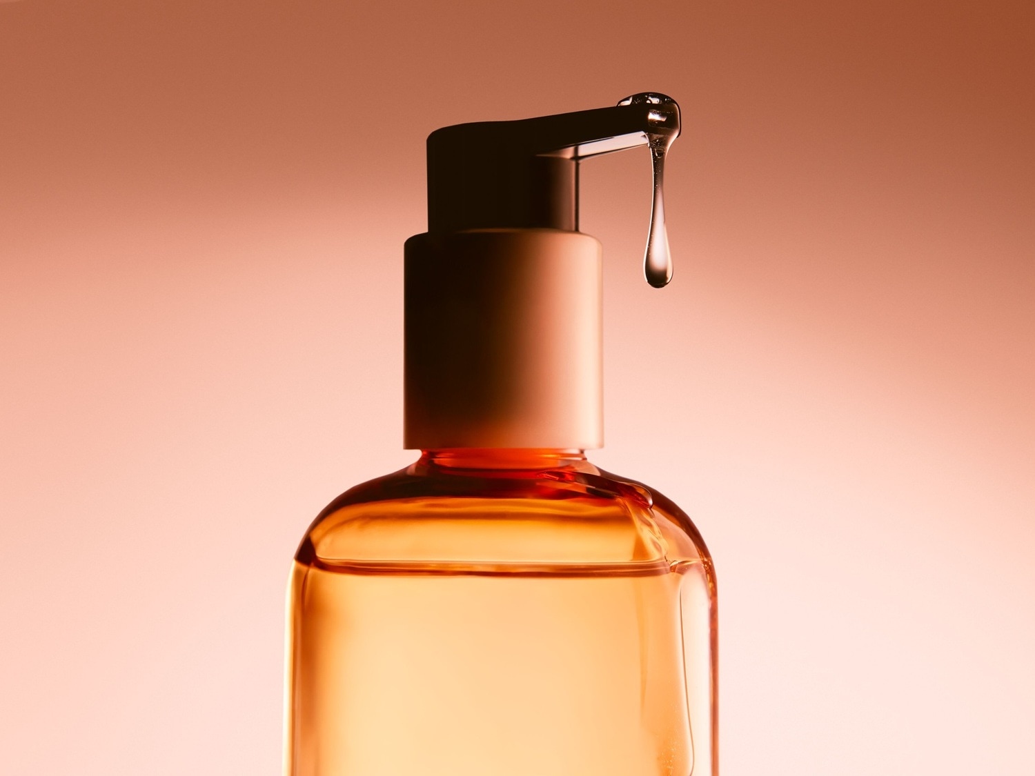 Nahaufnahme einer bernsteinfarbenen Flasche mit einer Pumpe, aus der ein Tropfen Serum austritt, vor einem sanften, pfirsichfarbenen Hintergrund.