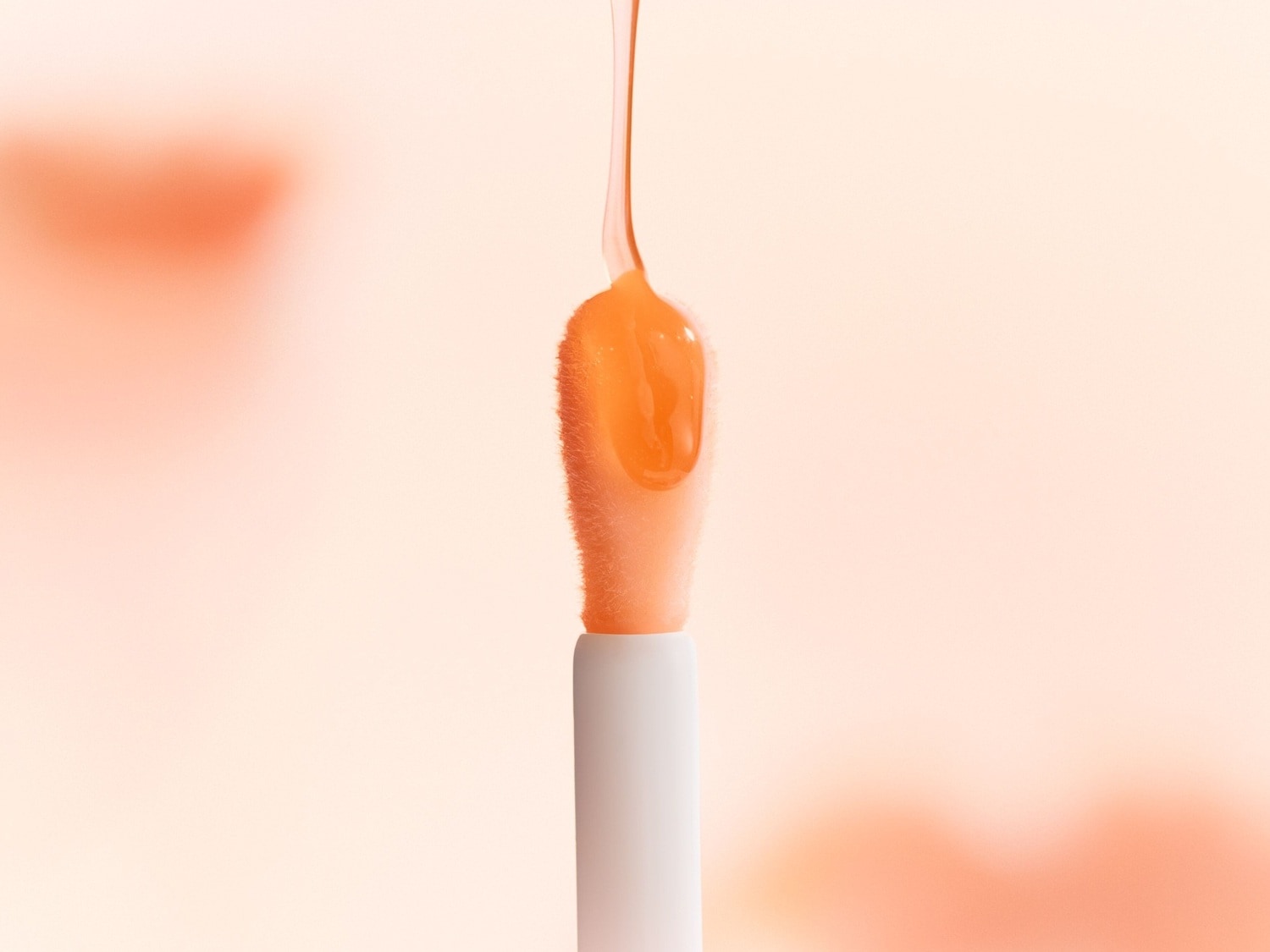 Nahaufnahme eines Applikators mit einem Tropfen glänzendem, orangefarbenem Lipgloss, der sich von einem hellen, pfirsichfarbenen Hintergrund abhebt.