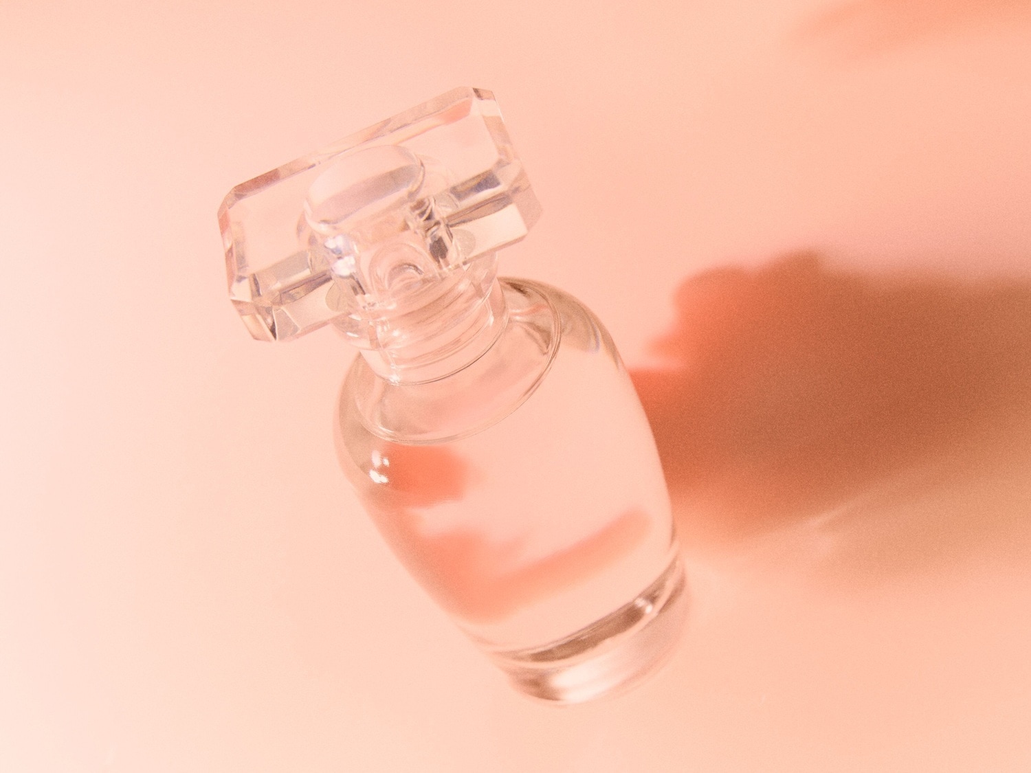 Ein durchsichtiger Glasflakon mit einem klaren, facettierten Verschluss, gefüllt mit einem hellen Parfüm, liegt auf einem sanft pfirsichfarbenen Hintergrund.