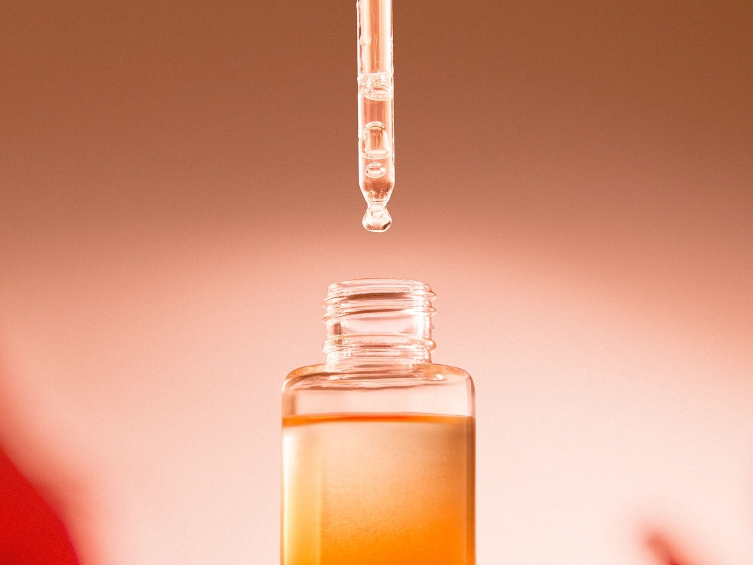 Nahaufnahme einer klaren Glasflasche mit goldfarbenem Serum und einem Applikator, der einen Tropfen abgibt, vor einem sanften, pfirsichfarbenen Hintergrund.