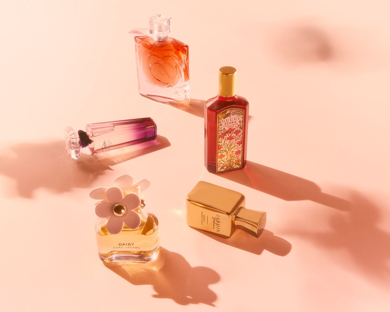 Eine Sammlung von Parfümflaschen verschiedener Marken und Düfte, darunter Daisy von Marc Jacobs und Gucci Flora, auf einem sanften, pfirsichfarbenen Hintergrund.