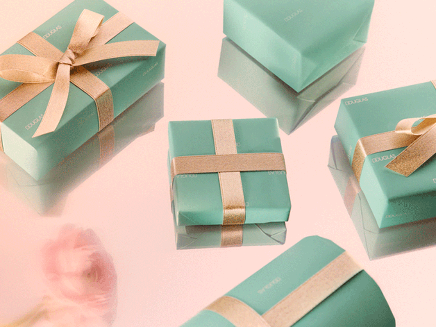 Elegantiškos dovanos žalios spalvos įvyniojime, papuoštos auksiniais kaspinais, išdėstytos ant švelniai rožinio fono, sukuria prabangos ir šventės atmosferą.