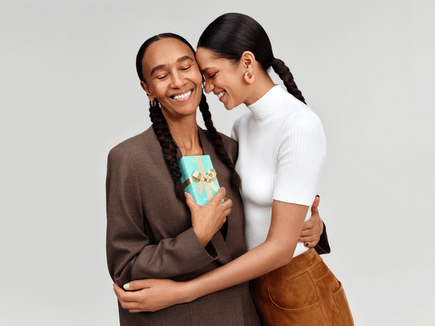 Švelnus mamos ir dukters apkabinimas, mama laiko mažą žydrą dovanų dėžutę su auksiniu kaspinu, abi šypsosi užmerktomis akimis.