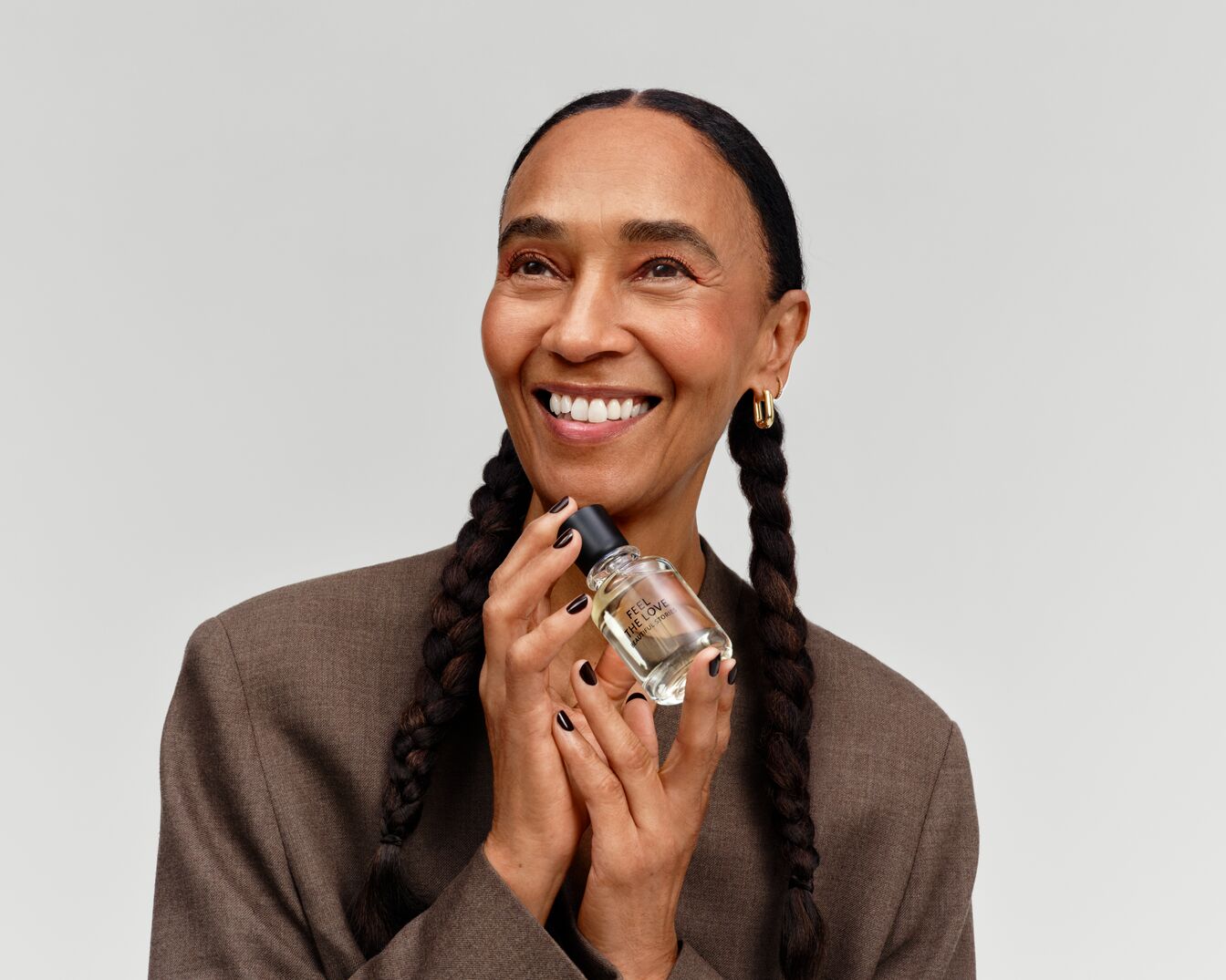 Eine lächelnde Frau mit Zöpfen hält eine durchsichtige Parfümflasche mit schwarzem Deckel in den Händen, auf der der Text "Feel the Love" steht.