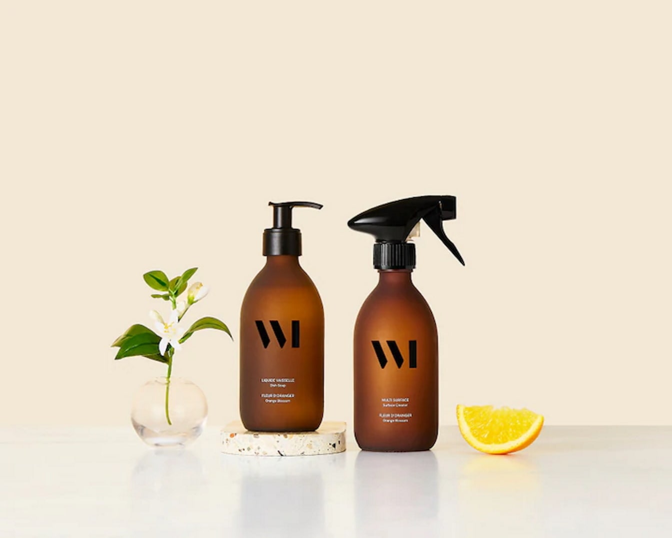 Deux bouteilles brunes avec étiquettes noires, une avec une pompe et l'autre avec un pulvérisateur, sont présentées avec une fleur blanche dans un vase et une tranche d'orange.