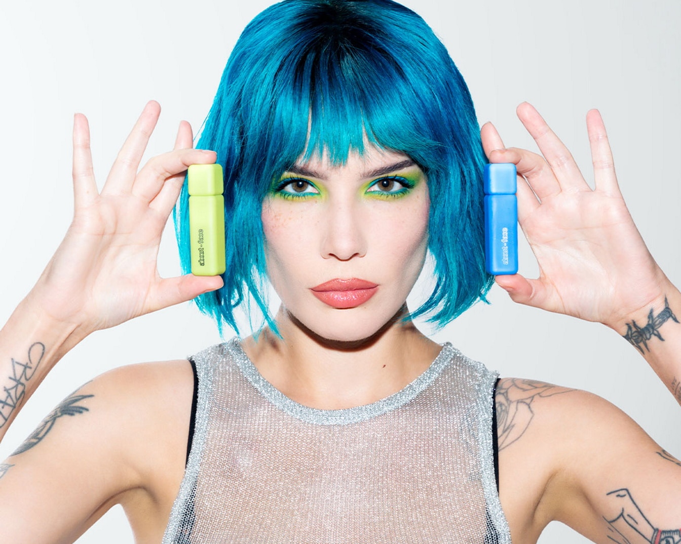 Une femme aux cheveux bleu turquoise présente deux flacons de parfum : un vert clair et un bleu vif, pour une expérience olfactive audacieuse et colorée.