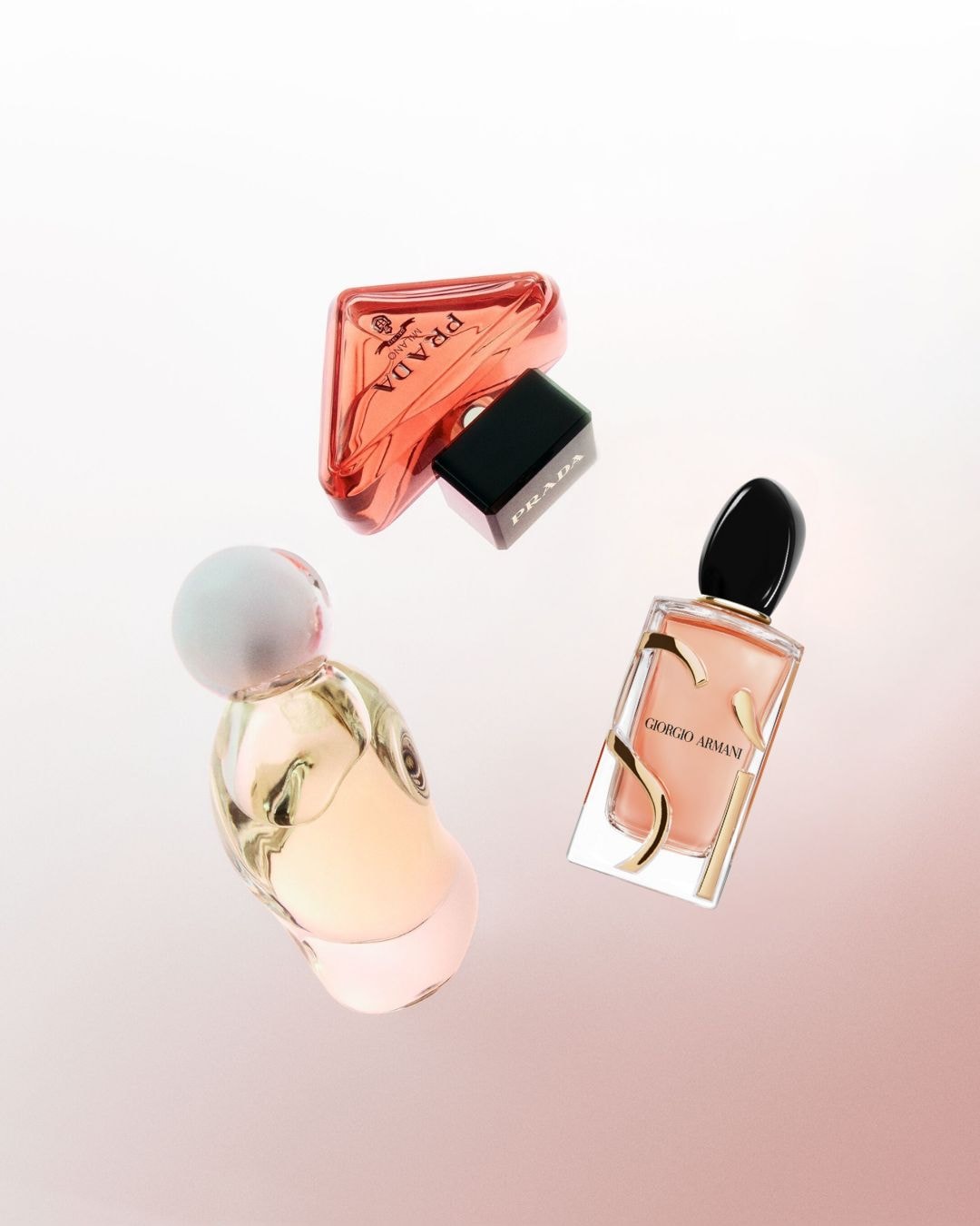 Drie parfumflesjes van Prada en Giorgio Armani zweven tegen een zachtroze achtergrond, elk met een unieke vorm en kleur.