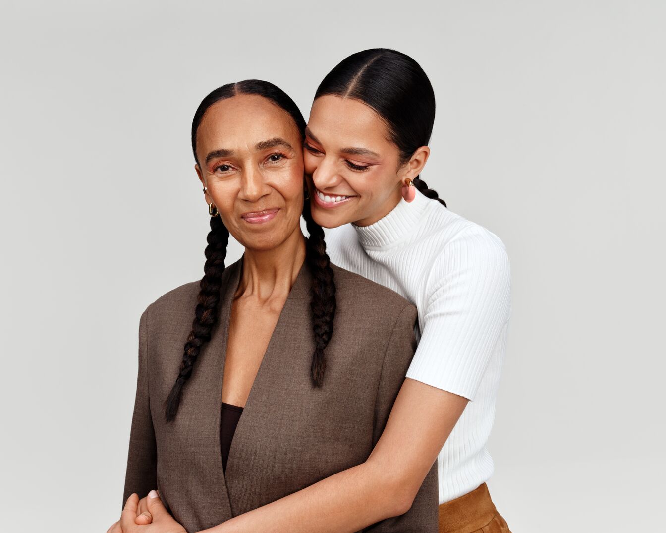 Eine lächelnde junge Frau umarmt liebevoll ihre Mutter, beide mit geflochtenen Zöpfen, die Mutter trägt einen braunen Blazer, während die Tochter einen weißen Rollkragenpullover trägt.