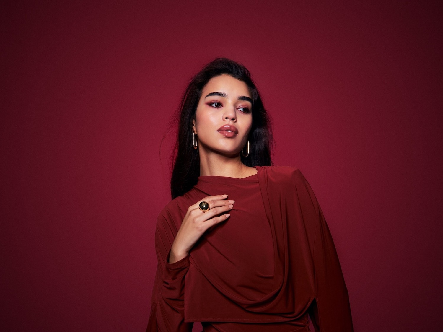 Une femme élégante avec un maquillage bordeaux et des lèvres brillantes, portant une robe bordeaux et des bijoux dorés, pose sur un fond bordeaux assorti.