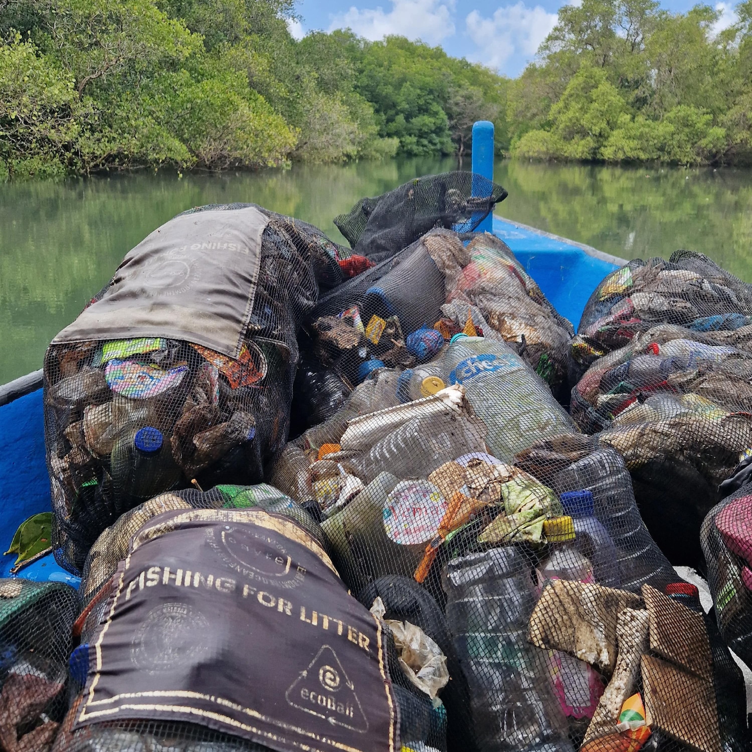 Una barca blu piena di sacchi neri con la scritta 'FISHING FOR LITTER' naviga su un fiume verde circondato da alberi, evidenziando l'iniziativa di pulizia ambientale.