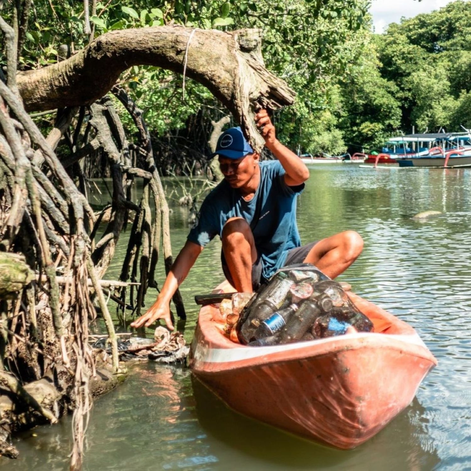 Un uomo su una canoa arancione raccoglie rifiuti da un fiume circondato da alberi, promuovendo la pulizia ambientale.