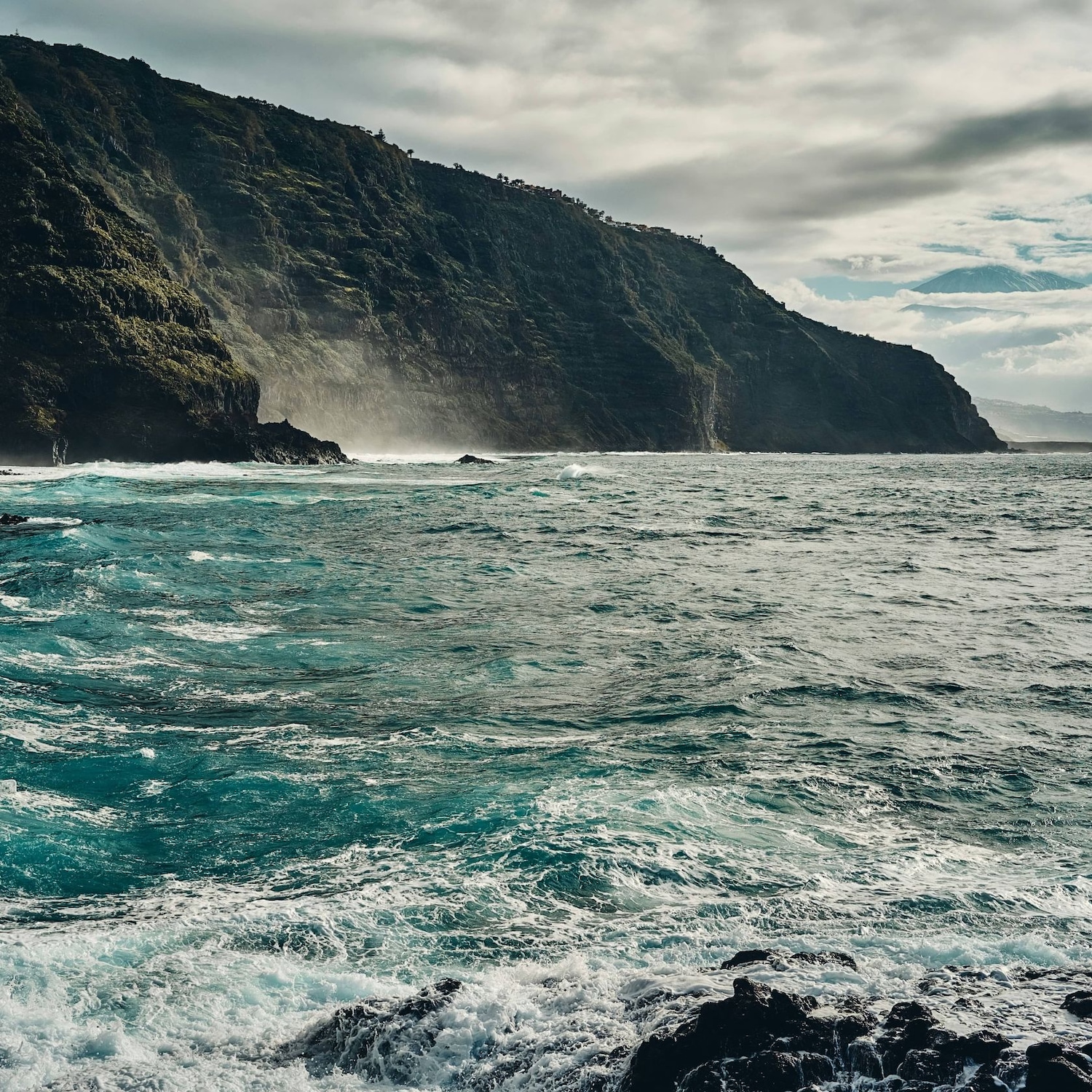 Una costa frastagliata incontra un mare mosso sotto un cielo nuvoloso, con una montagna verdeggiante che si erge sullo sfondo.
