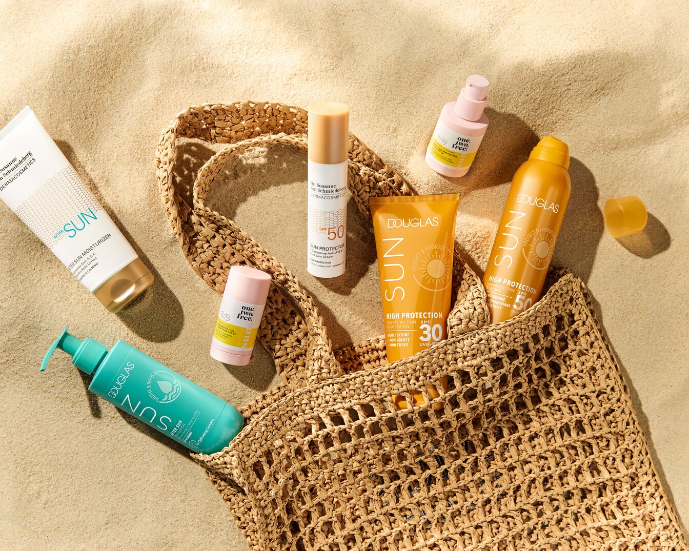 Verschiedene Sonnenschutzprodukte wie Lotionen und Sprays von Marken wie Douglas und One.Two.Free liegen auf einem Sandstrand neben einer Strandtasche.