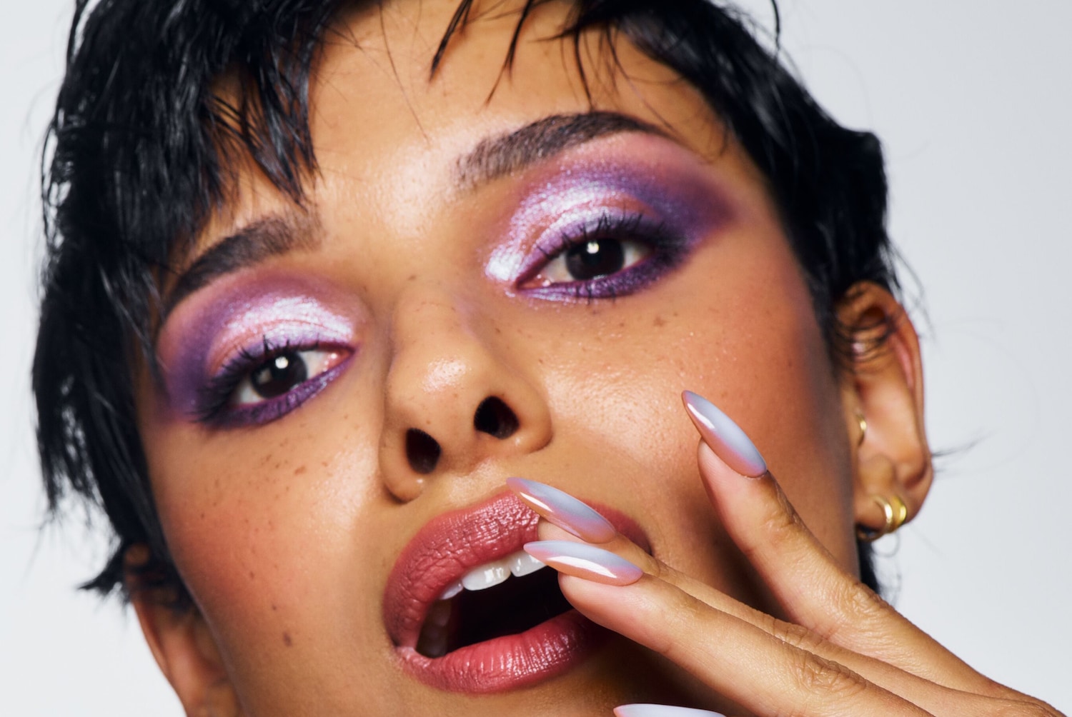 Nahaufnahme eines Gesichtes mit auffälligem lila und silbernem Lidschatten, glänzenden Lippen und mandelförmigen Nägeln, die in einem sanften Farbverlauf von Rosa zu Weiß übergehen.