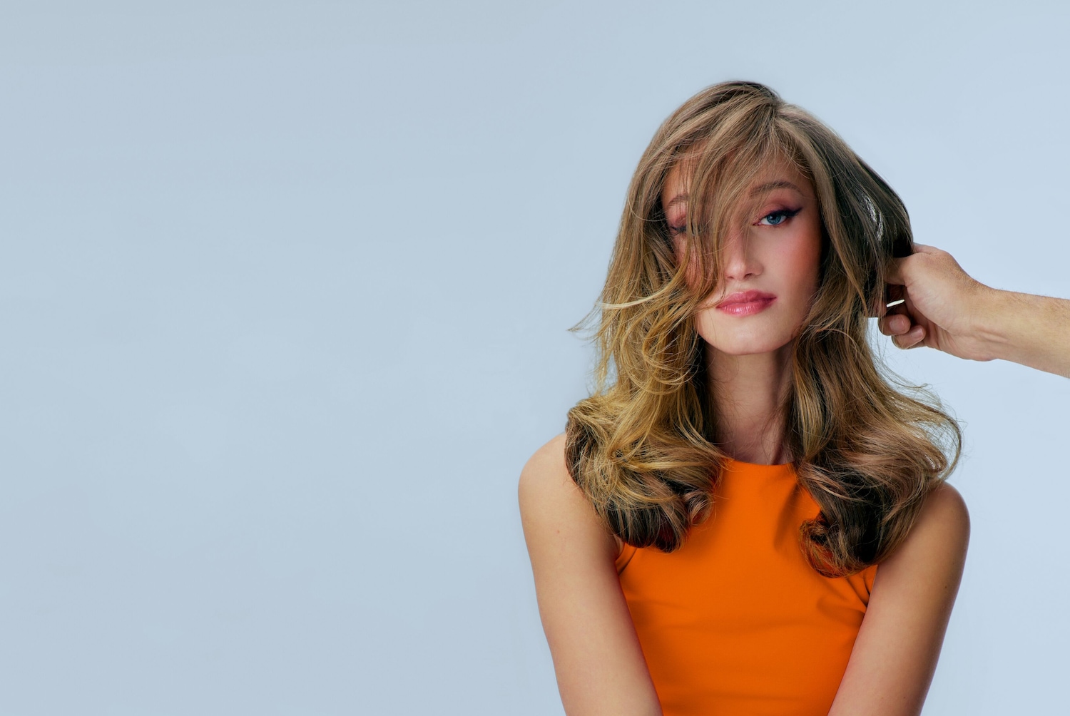 Eine Frau mit gewelltem, blondem Haar und einem orangefarbenen Oberteil posiert, während eine Hand ihr Haar berührt.