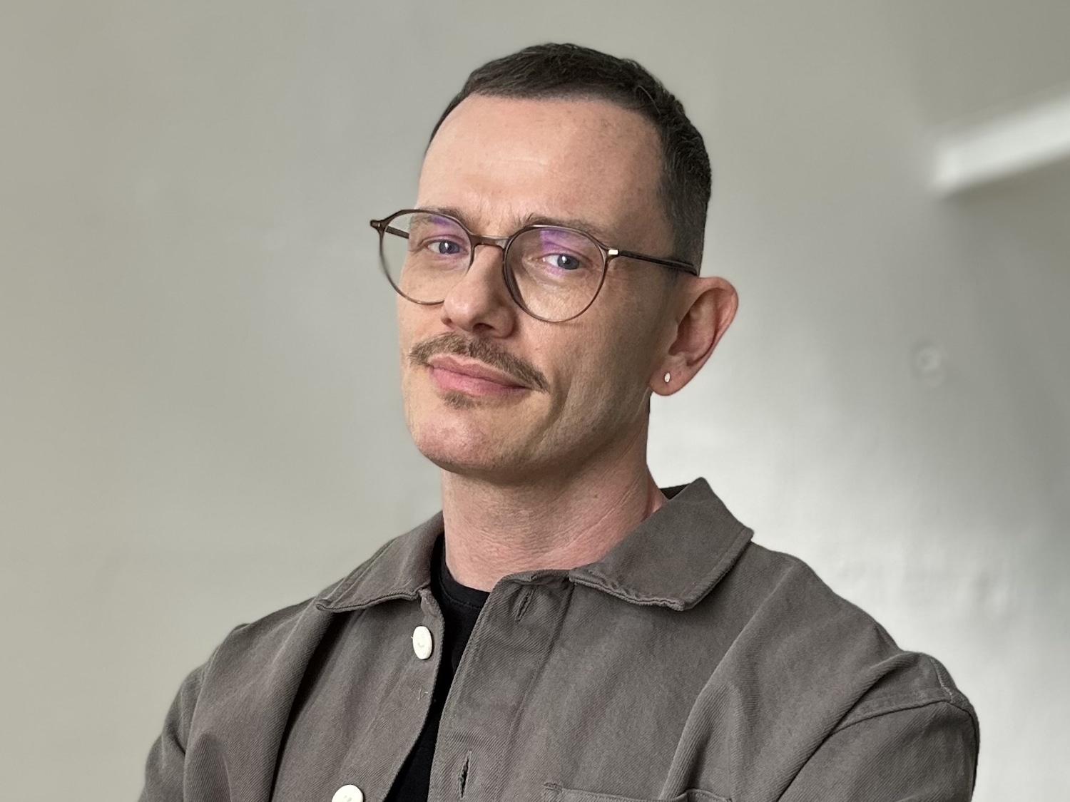 Porträt eines Mannes mit Brille, Schnurrbart und grauem Hemd vor einem hellen Hintergrund.